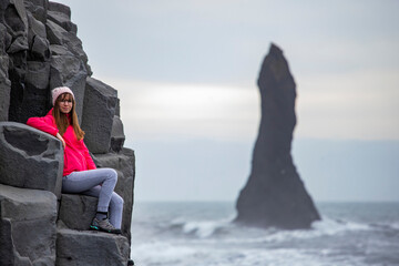 Beautiful girl in pink jacket sits on basalt columns with big rock (troll fingers) on famous Reynisfjara black beach, Iceland