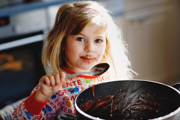 Cute toddler girl eating chocolate dough rests with spoon and fingers from pot. Happy child licking sweet dough for muffins or cake, helping in home kitchen, indoors.