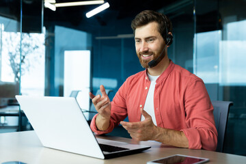 Learning online. A young male teacher in a headset and a red shirt sits in the office at a table, works on a laptop, teaches through a video call, conference, webinar.