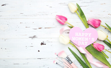 Canvas Print - Flowers of tulips, card in shape of heart with text Happy Women's Day, pink makeup brushes and ribbon on white wooden background