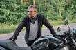 © perfectlab - Man biker middle aged motorcyclist smiling stands with a motorcycle on the side of the road in the forest