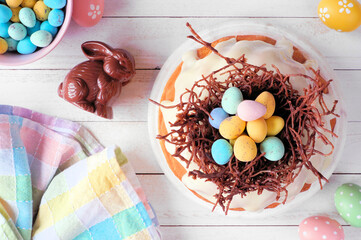 Poster - Easter bundt cake with chocolate nest of colorful candy eggs. Overhead view table scene on a white wood background.