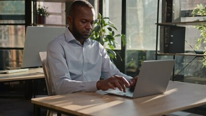 Focused adult senior businessman mature entrepreneur African American ethnic man working in office business firm programming on computer keyboard online project typing email sit in sunbeams background