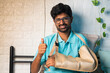 © WESTOCK - happy smiling young man with broken hand at bedroom showing thumbs up gesture by looming at camera - concept of recovered, successful medical treatment and rehabilitation