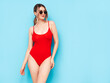 © halayalex - Young beautiful sexy woman. Smiling carefree model wearing red lingerie. Hot tanned blond posing near blue wall in studio in summer swimwear bathing suit. Cheerful and happy. In sunglasses