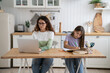 © DimaBerlin - Concentrated woman tutor sits at kitchen table with teenage girl providing extracurricular services. Busy casual mother with laptop and diligent school age daughter with books and workbook. Freelance.