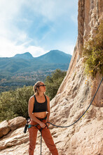 A Rock Climber Stands In A Canyon, Holding A Rope. The Climber Insures His Partner. Sports Girl Smiles And Holds Climbing Equipment. Climber Near The Cliff.