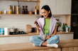 © Drobot Dean - Smiling asian woman using mobile phone and drinking coffee while sitting in kitchen