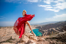 Woman Red Dress Lake Mountains. Side View Of A Woman In A Long Red Dress Posing On A Rock High Above The Lake. Against The Background Of The Blue Sky And The Lake In The Form Of A Heart