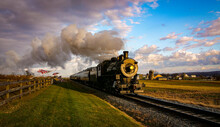 A View Of A Classic Steam Passenger Train Approaching, With American Flags Attached To A Fence On A Sunny Autumn Day