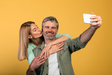 Wall Mural - Happy middle aged wife kissing husband on cheek, spouses making selfie on yellow background, studio shot