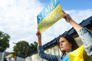 Poster - Sad woman holding poster in colors of national flag and words Stand with Ukraine outdoors. Space for text