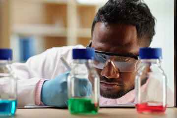 Wall Mural - Serious scientist in protective glasses looking at bottles with reagents and taking notes in notebook