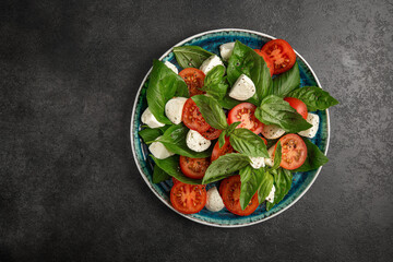 Wall Mural - Fresh italian caprese salad with red tomato, green leaf basil, mozzarella cheese on a dark stone table.