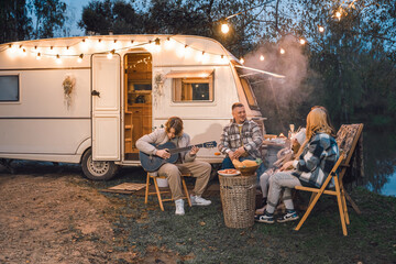 Wall Mural - Family trailer travel.Children,brother sister,mom dad playing guitar,singing song at fire.Evening picnic in nature.Holiday barbecue BBQ food.Vacation weekend dinner.Road lunch.Camper,house on wheels