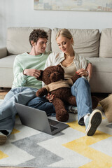 Wall Mural - cheerful young woman and happy man holding teddy bear near laptop in living room.