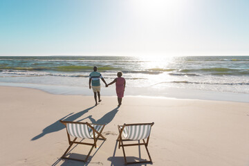 Rear view of african american senior couple holding hands and walking towards sea under sky