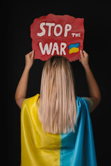 Wall Mural - Woman wrapped in Ukrainian flag and holding poster with words Stop the War on black background, back view