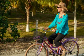 Front view closeup of happy smiling pretty mature senior woman smiling biking towards the camera wearing ethnic clothes and hat and sunglasses on a tropical road.