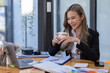 © David - Beautiful young smiling Asian businesswoman working on laptop and drinking coffee, Asia businesswoman working document finance and calculator in her office.