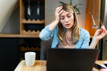 A tired woman holds her head while holding glasses in her hand. She is tired of working overtime