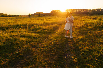 Wall Mural - Wide shot of loving young couple in relationship hugging and kissing enjoying each other in wonderful sunlight of sunset on green meadow. Happy man and woman spending unforgettable moments together.