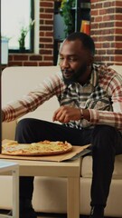 Wall Mural - Vertical video: Happy guy eating slice of pizza and drinking beer at home, enjoying takeout food and watching television. Young man serving takeaway dinner and alcohol, watch movie in living room.