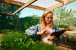 © StratfordProductions - Young caucasian woman in checked shirt pointing at digital tablet by plants in plant nursery