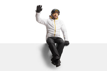 Sticker - Young man wearing a skiing equipment sitting on a blank panel and waving