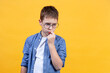 Portrait of a boy with glasses looking right and down. He is thinking, frowned, and put a finger to his lip. Yellow background. Front view. Copy space