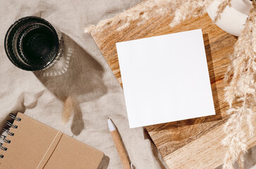 Blank paper sheet card mockup on wooden desk, vase with dried lagurus grass, notepad and glass of water with sunlight shadows on beige linen cloth. Minimal aesthetic brand template, home office desk