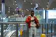 Young smiling african man traveler with luggage texting sms message to family or friends via mobile phone after arrival in airport, happy black guy with smartphone in terminal, booking hotel or taxi