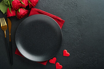 Valentines day romantic table setting. Empty closeup of red roses, wine, candles, dinner black plate, knife, fork and decorative silk hearts on black background. Holiday concept. Copy space. Top view