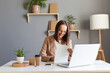 © sementsova321 - Indoor shot of smiling young adult beautiful brown haired woman wearing beige jacket posing in office, working on laptop and and with paper documents, enjoying her work.
