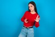 Joyful young brunette girl wearing red T-shirt against blue wall wink and points index fingers at camera, chooses someone, makes finger gun pistol.
