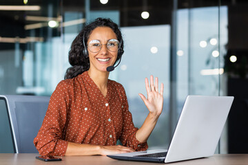 Portrait of successful tech support worker, hispanic woman with curly hair smiling and looking at camera, businesswoman with headset for video call, greeting gesture, inside office with laptop.