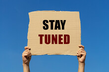 Stay Tuned Text On Box Paper Held By 2 Hands With Isolated Blue Sky Background. This Message Board Can Be Used For Business Concept About Being Ready For Any Event That Will Be Started Soon.