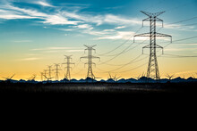 Sunset Silhouette Of Steel Lattice Transmission Towers And Power Lines Overlooking Distant Mountains And Windmills.