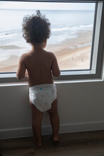 Cute Boy Toddler On Summer Vacation Looking Out Of The Hotel Window At The Beach And Ocean. He Has Brown Curly Hair And Is Wearing A Diaper As Seen From Back.