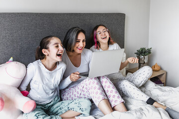 Wall Mural - Hispanic women family enjoying live stream or watching a movie with computer or laptop on bed together mother and daughters in Latin America	