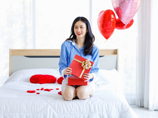 Millennial Asian young romantic female girlfriend sitting on bed in bedroom decorate with pink heart shape balloon holding opening red gold ribbon wrapped surprise gift box on valentine day festive