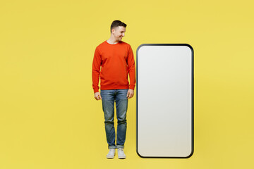 Wall Mural - Full body smiling young man wear orange casual clothes stand near big huge blank screen mobile cell phone looking at smartphone with copy space mockup area isolated on plain yellow color background.