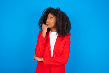 Wall Mural - Portrait of thoughtful young businesswoman with afro hairstyle wearing red over blue, keeps hand under chin, looks away trying to remember something or listens something with interest. Youth concept.