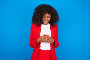 Wall Mural - Smiling young businesswoman with afro hairstyle wearing red over blue wall using cell phone, messaging, being happy to text with friends, looking at smartphone. Modern technologies and communication.