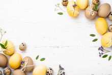 Festive Easter Background. Yellow And Brown Easter Eggs With Flowers On A White Wooden Table. Greeting Card With Place For Text. Top View.