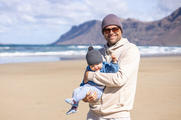 Wall Mural - Father enjoying pure nature holding and playing with his infant baby boy son in on windy sandy beach of Famara, Lanzarote island, Spain. Family travel and parenting concept