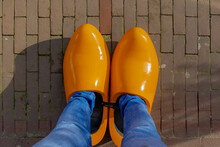 Top View Of A Man With Blue Jeans Standing In Orange Wooden Shoes On The Bricks Ground Floor, Low Angle Of A Man Wearing Oversized Or Giant Clogs (Dutch Wooden Shoes) Netherlands.