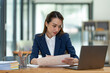 © crizzystudio - Asian businesswoman sitting happily in office holding detailed report paper analyzing data accuracy in financial matters management concept.