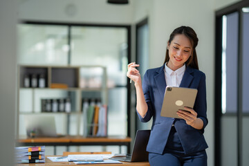 Asian businesswoman in the suit standing confidently holding iPad or tablet in hand and smiling in the office.