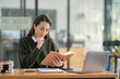 © crizzystudio - Beautiful Asian businesswoman working on a laptop and having fun at work and reading review book documenting detailed financial information earning peace of mind.